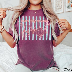 May include: Berry-colored t-shirt with a graphic design featuring light blue vertical stripes and beach-themed illustrations. The text "wanna play mermaid?" is printed across the design. The shirt has a relaxed fit.