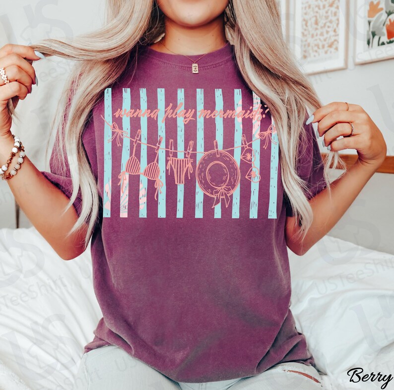 May include: Berry-colored t-shirt with a graphic design featuring light blue vertical stripes and beach-themed illustrations. The text "wanna play mermaid?" is printed across the design. The shirt has a relaxed fit.
