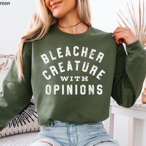 Puede incluir: Una sudadera verde militar con el texto "BLEACHER CREATURE WITH OPINIONS" en blanco. La sudadera es de estilo cuello redondo y manga larga. La imagen muestra la parte superior del cuerpo de una persona que lleva la sudadera.