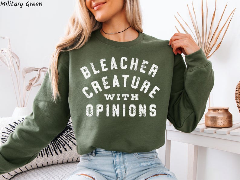 Puede incluir: Una sudadera verde militar con el texto "BLEACHER CREATURE WITH OPINIONS" en blanco. La sudadera es de estilo cuello redondo y manga larga. La imagen muestra la parte superior del cuerpo de una persona que lleva la sudadera.
