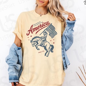 Puede incluir: Camiseta de color mantequilla con un dise&ntilde;o patri&oacute;tico. El gr&aacute;fico incluye la palabra "America" en rojo, un jinete a caballo sosteniendo una bandera estadounidense y el texto "Since 1776". La camiseta tiene mangas cortas y un corte relajado.