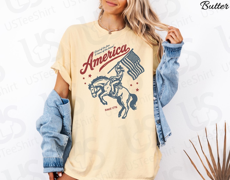Puede incluir: Camiseta de color mantequilla con un dise&ntilde;o patri&oacute;tico. El gr&aacute;fico incluye la palabra "America" en rojo, un jinete a caballo sosteniendo una bandera estadounidense y el texto "Since 1776". La camiseta tiene mangas cortas y un corte relajado.