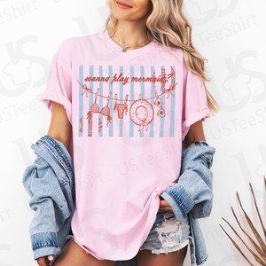 May include: Pink t-shirt with a graphic of a bikini, swim trunks, a sun hat, and the text "wanna play mermaids?" on a blue and white striped background. The shirt is worn by a person.
