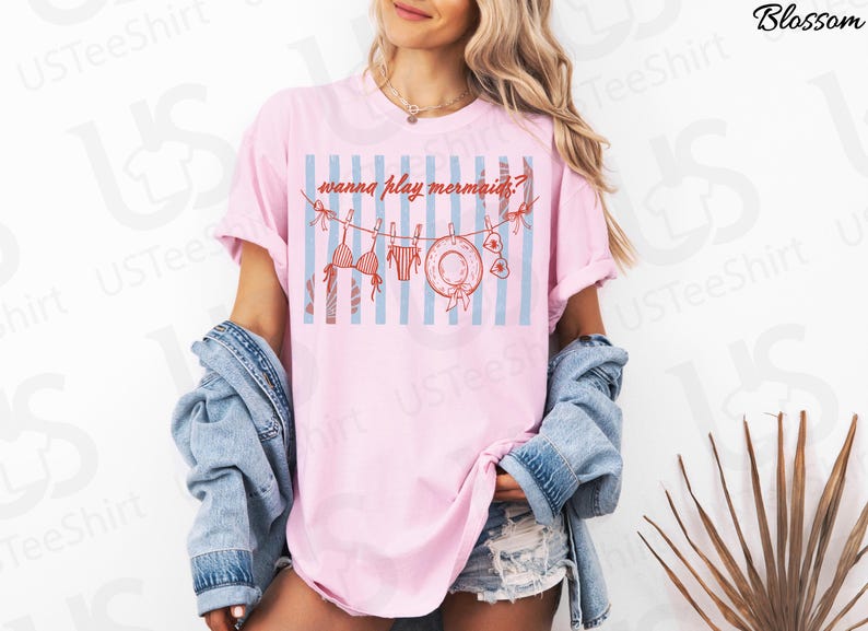 May include: Pink t-shirt with a graphic of a bikini, swim trunks, a sun hat, and the text "wanna play mermaids?" on a blue and white striped background. The shirt is worn by a person.