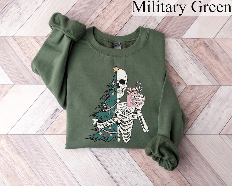 May include: A military green crewneck sweatshirt featuring a graphic print of a split figure that is half Christmas tree and half skeleton. The skeleton holds a pink mug with candy canes. A banner across the center reads "SORTA MERRY SORTA SCARY".