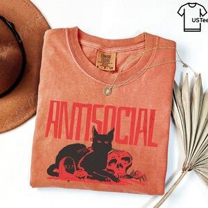 K&ouml;nnte beinhalten: Orangefarbenes Comfort Colors T-Shirt mit dem Wort "ANTISOCIAL" in roten Blockbuchstaben. Eine schwarze Katze, ein Totenkopf, ein Grabstein und Spinnen sind unter dem Text abgebildet. Eine goldene Halskette und ein brauner Hut sind ebenfalls im Bild.