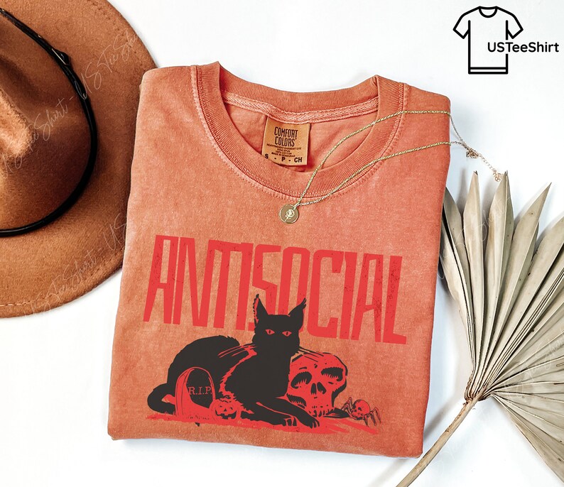 K&ouml;nnte beinhalten: Orangefarbenes Comfort Colors T-Shirt mit dem Wort "ANTISOCIAL" in roten Blockbuchstaben. Eine schwarze Katze, ein Totenkopf, ein Grabstein und Spinnen sind unter dem Text abgebildet. Eine goldene Halskette und ein brauner Hut sind ebenfalls im Bild.