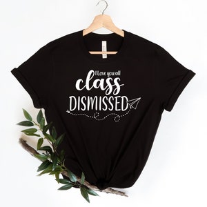 May include: Black t-shirt with the white text "I love you all class DISMISSED" and a paper airplane graphic. The shirt is hanging on a wooden hanger. A sprig of greenery is placed on the lower left side of the shirt.