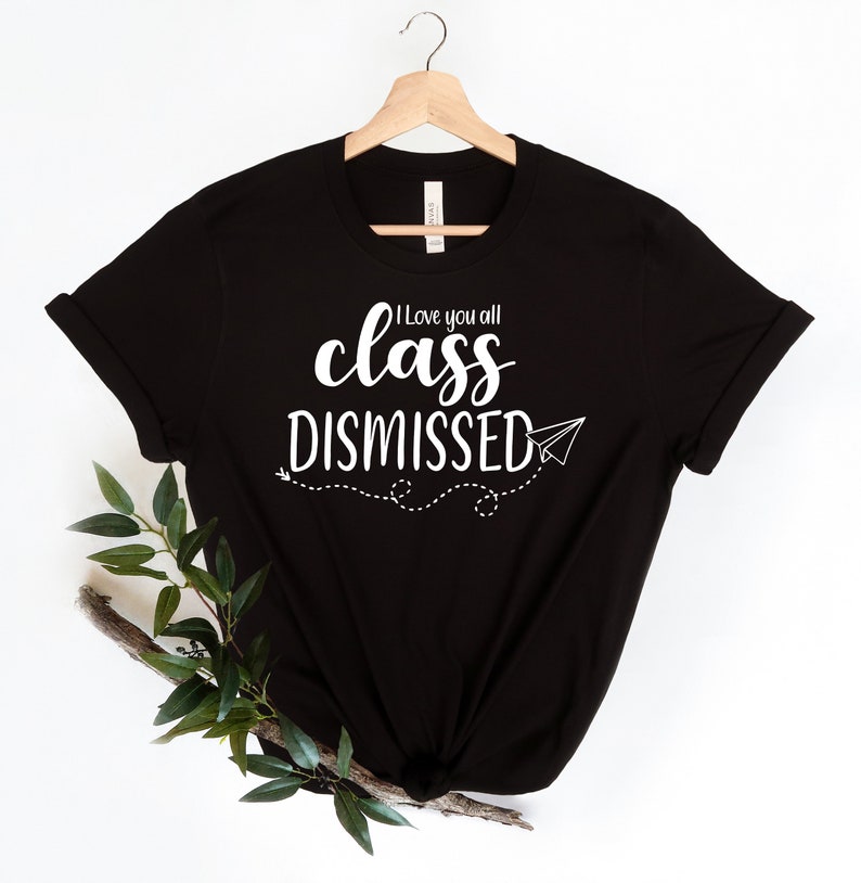 May include: Black t-shirt with the white text "I love you all class DISMISSED" and a paper airplane graphic. The shirt is hanging on a wooden hanger. A sprig of greenery is placed on the lower left side of the shirt.