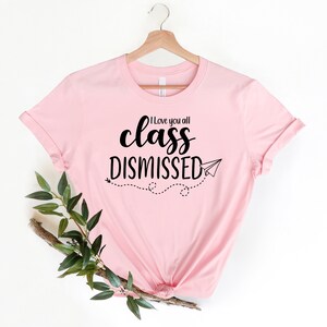 May include: A light pink t-shirt with the words "I love you all class DISMISSED" in black script and a paper airplane graphic. The shirt is displayed on a wooden hanger.