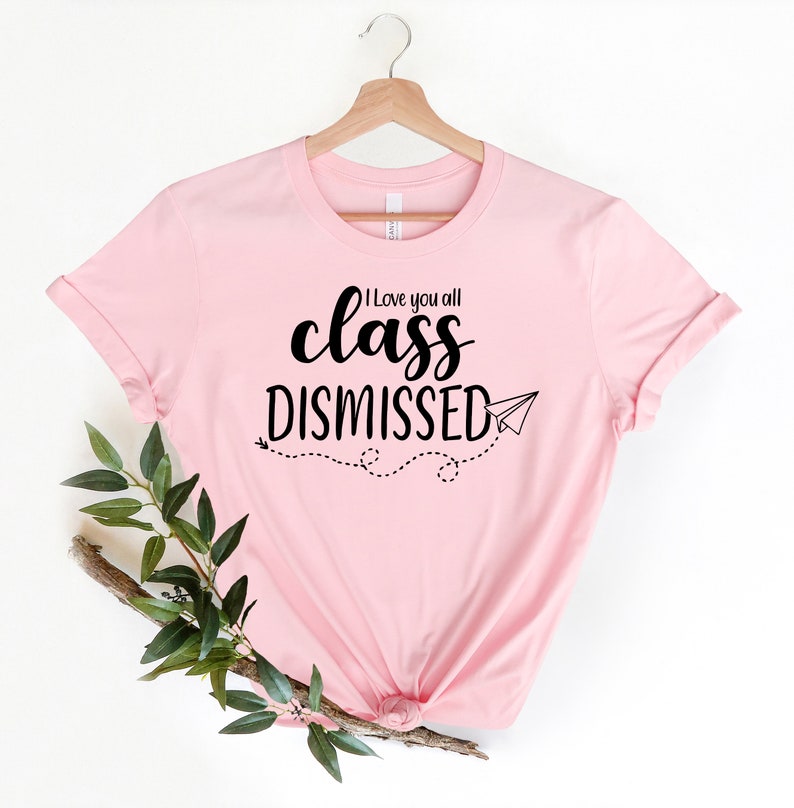 May include: A light pink t-shirt with the words "I love you all class DISMISSED" in black script and a paper airplane graphic. The shirt is displayed on a wooden hanger.