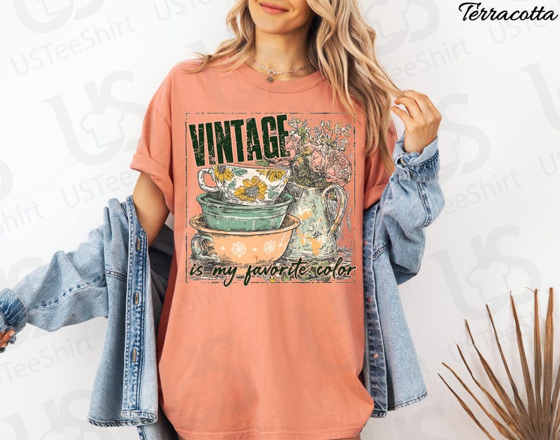 May include: Terracotta-colored t-shirt with a vintage-style graphic. The graphic features stacked teacups and bowls, a floral pitcher, and the text "VINTAGE is my favorite color" in a distressed font. The shirt is a relaxed fit.