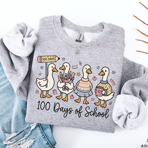 May include: A light gray sweatshirt with a whimsical graphic of four ducks celebrating 100 days of school. The ducks are holding school supplies and wearing sweaters. The text "100 Days of School" is written below the ducks.