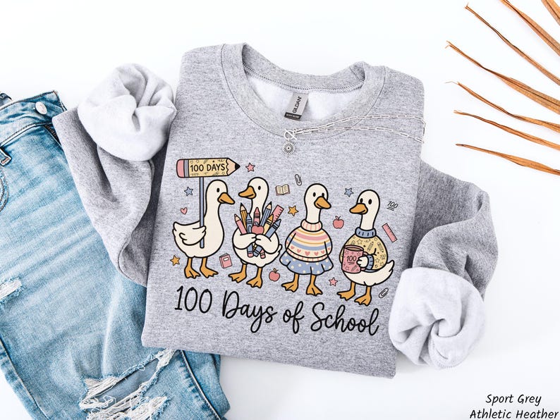 May include: A light gray sweatshirt with a whimsical graphic of four ducks celebrating 100 days of school. The ducks are holding school supplies and wearing sweaters. The text "100 Days of School" is written below the ducks.
