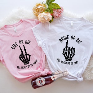 Puede incluir: Dos camisetas, una rosa y otra blanca, con texto y gr&aacute;ficos negros. La camiseta rosa dice "RIDE OR DIE" con una mano de esqueleto haciendo la se&ntilde;al de la paz. La camiseta blanca dice "BRIDE OR DIE" con una mano de esqueleto haciendo una peineta.