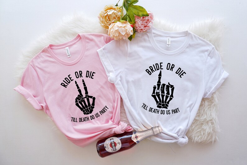 Puede incluir: Dos camisetas, una rosa y otra blanca, con texto y gr&aacute;ficos negros. La camiseta rosa dice "RIDE OR DIE" con una mano de esqueleto haciendo la se&ntilde;al de la paz. La camiseta blanca dice "BRIDE OR DIE" con una mano de esqueleto haciendo una peineta.
