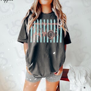 May include: Dark gray t-shirt with a distressed graphic featuring a hat, bikini, and the text "gonna have memories" in pink. The design includes vertical light blue stripes. The shirt is a relaxed fit.