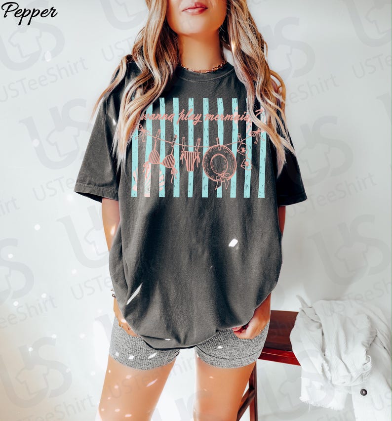 May include: Dark gray t-shirt with a distressed graphic featuring a hat, bikini, and the text "gonna have memories" in pink. The design includes vertical light blue stripes. The shirt is a relaxed fit.