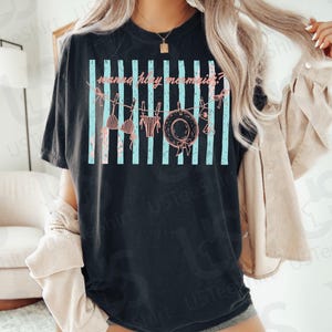 May include: Black t-shirt with a graphic that says "wanna play mermaids?" in pink script. The design includes a clothesline with a bikini top, bottoms, and a sun hat. Light blue vertical stripes are behind the clothesline.