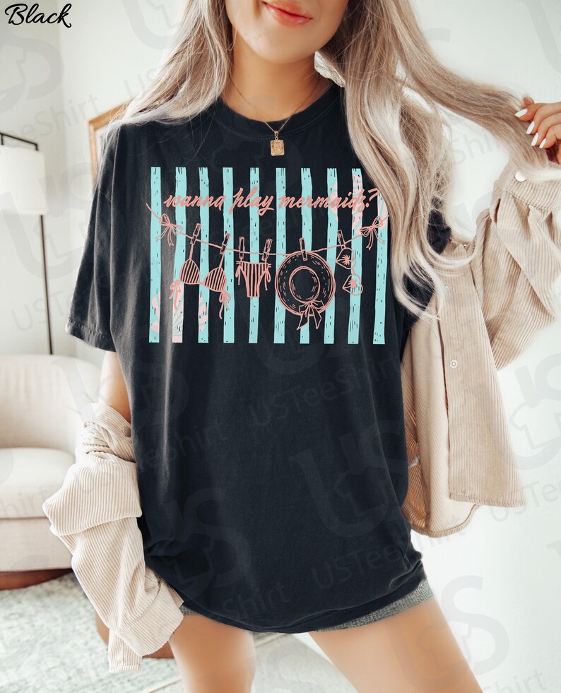 May include: Black t-shirt with a graphic that says "wanna play mermaids?" in pink script. The design includes a clothesline with a bikini top, bottoms, and a sun hat. Light blue vertical stripes are behind the clothesline.