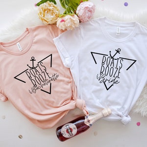 Op de afbeelding: Een perzikkleurig t-shirt met zwarte tekst "BOATS BOOZE & Bachelorette" en een wit t-shirt met zwarte tekst "BOATS BOOZE & Bride". Beide t-shirts hebben een zwart grafisch ontwerp met een anker en een driehoek.
