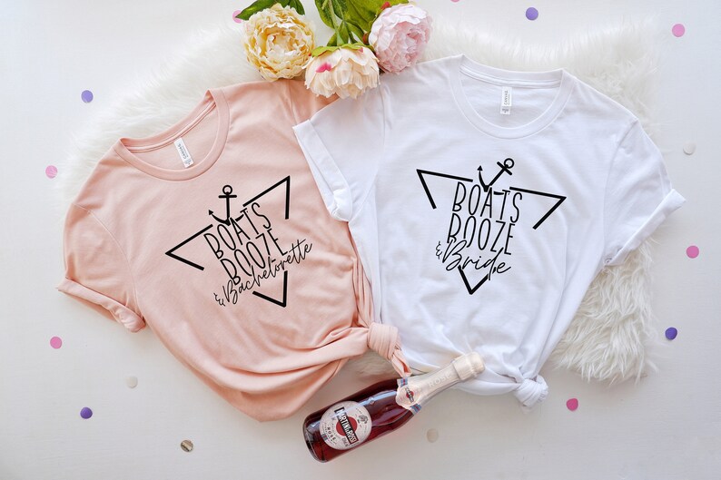 Op de afbeelding: Een perzikkleurig t-shirt met zwarte tekst "BOATS BOOZE & Bachelorette" en een wit t-shirt met zwarte tekst "BOATS BOOZE & Bride". Beide t-shirts hebben een zwart grafisch ontwerp met een anker en een driehoek.