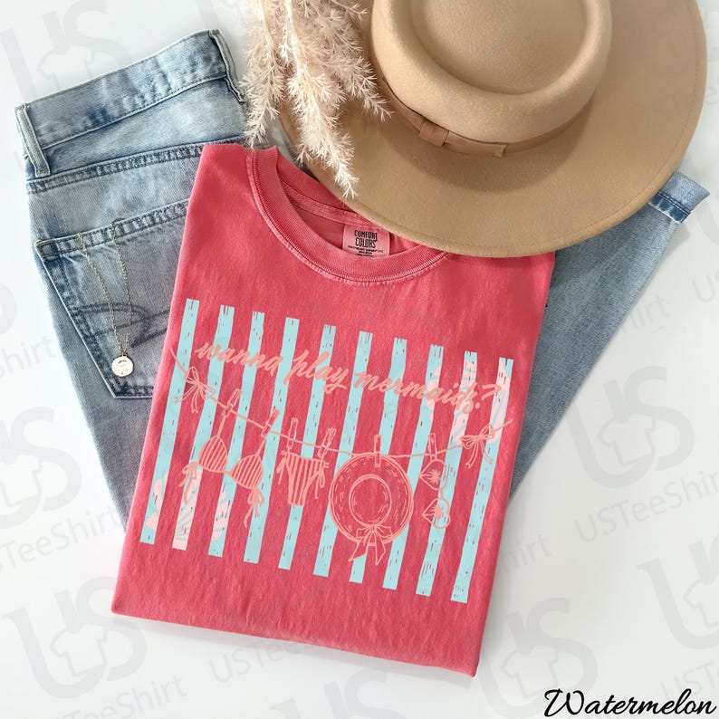 May include: A red t-shirt with the text "wanna holiday mermaid?" in a light blue font, with a beach theme design. The shirt is paired with light blue jeans and a tan hat.