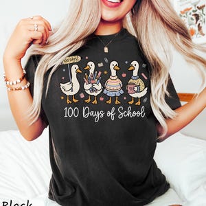 May include: Black t-shirt with a whimsical graphic of five ducks celebrating "100 Days of School." Each duck is uniquely styled, holding school supplies and wearing sweaters. The text "100 Days of School" is written in a cursive font.