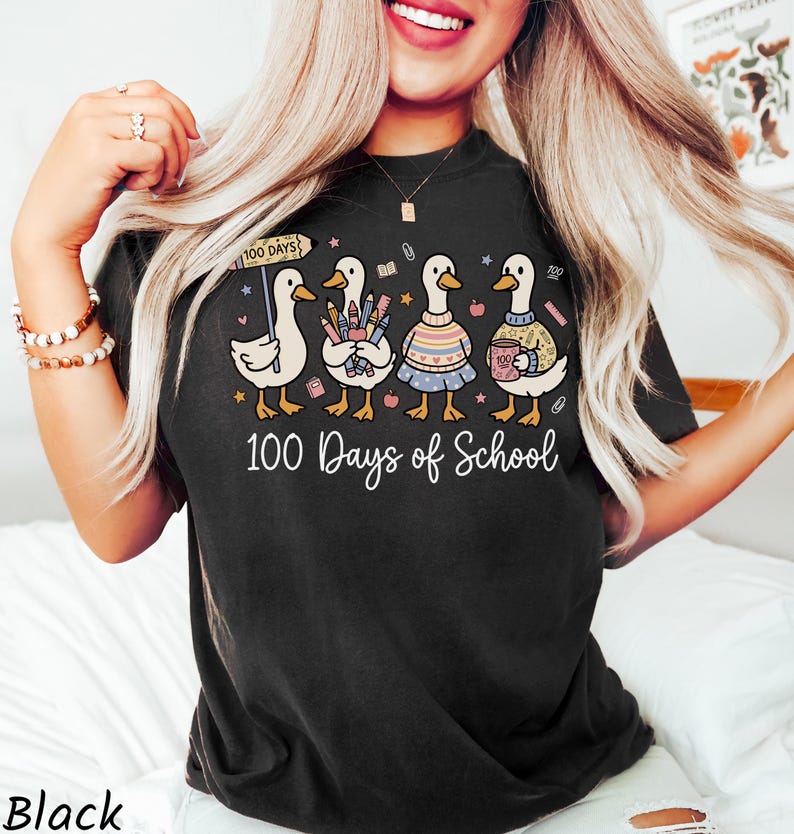 May include: Black t-shirt with a whimsical graphic of five ducks celebrating "100 Days of School." Each duck is uniquely styled, holding school supplies and wearing sweaters. The text "100 Days of School" is written in a cursive font.