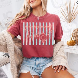 May include: Crimson-colored t-shirt with a graphic design featuring a beach theme. The design includes vertical light blue stripes, a hat, and swimwear. The text on the shirt reads "mermaid holiday?"
