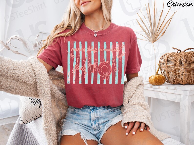 May include: Crimson-colored t-shirt with a graphic design featuring a beach theme. The design includes vertical light blue stripes, a hat, and swimwear. The text on the shirt reads "mermaid holiday?"
