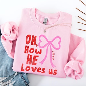 May include: A light pink sweatshirt with the phrase "OH, How HE Loves us" in red text and a pink bow graphic. A delicate necklace with a small pendant rests on the sweatshirt. The sweatshirt is folded, with a pair of blue jeans visible.