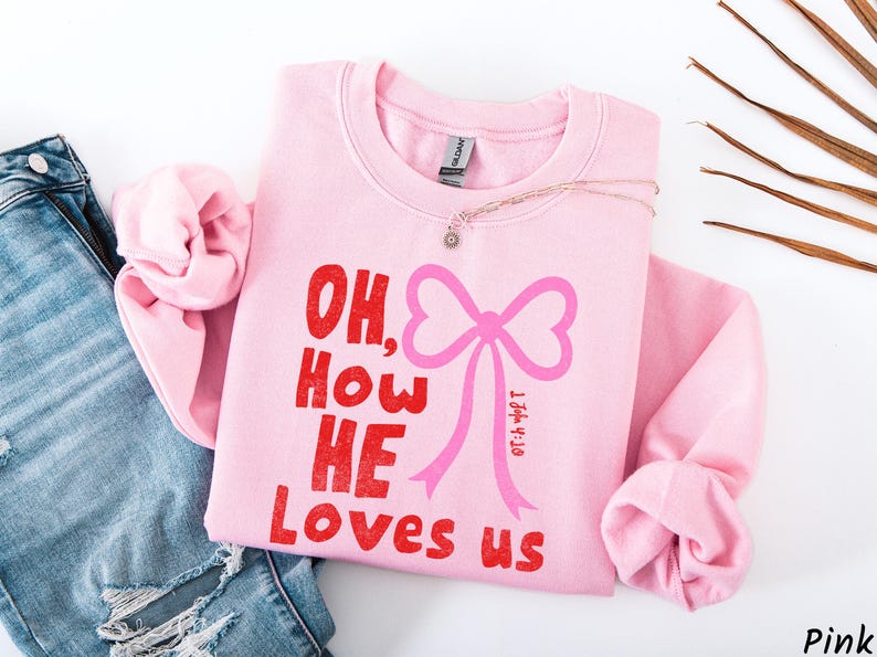 May include: A light pink sweatshirt with the phrase "OH, How HE Loves us" in red text and a pink bow graphic. A delicate necklace with a small pendant rests on the sweatshirt. The sweatshirt is folded, with a pair of blue jeans visible.