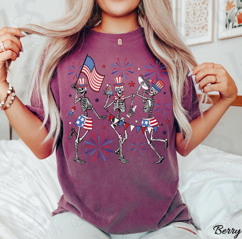 May include: Berry-colored t-shirt featuring a festive design of dancing skeletons celebrating with American flags, cupcakes, and fireworks. The skeletons wear patriotic hats and skirts, creating a fun and celebratory theme.