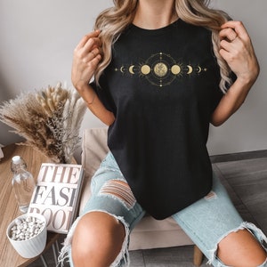 May include: Black t-shirt featuring a gold celestial design with moon phases and a central full moon. The design is intricate and detailed, with a geometric arrangement of stars and lines. The shirt is a classic crew neck style.
