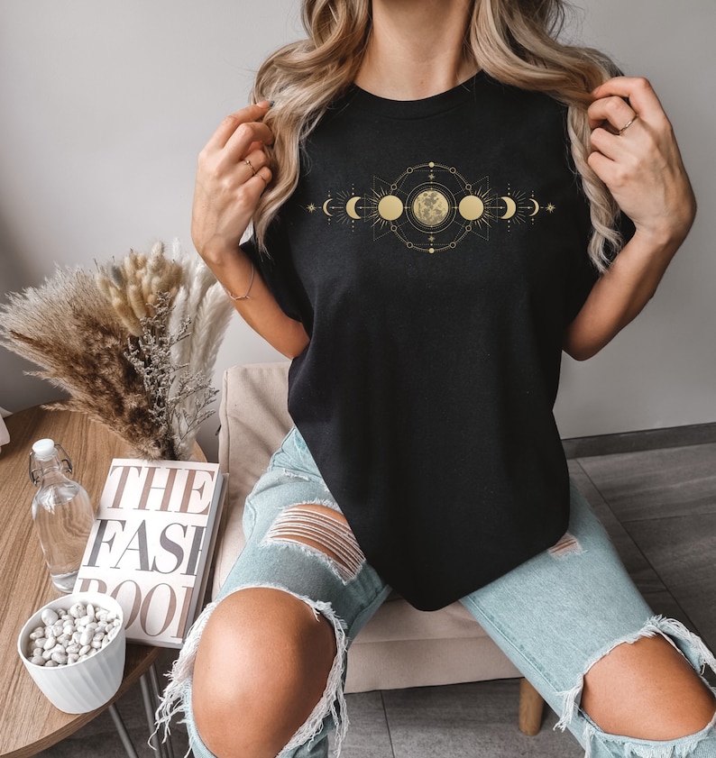 May include: Black t-shirt featuring a gold celestial design with moon phases and a central full moon. The design is intricate and detailed, with a geometric arrangement of stars and lines. The shirt is a classic crew neck style.