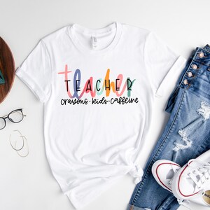 May include: White t-shirt with the word "TEACHER" in colorful letters, with "crayons, kids, caffeine" below. The shirt is paired with blue jeans and white sneakers.
