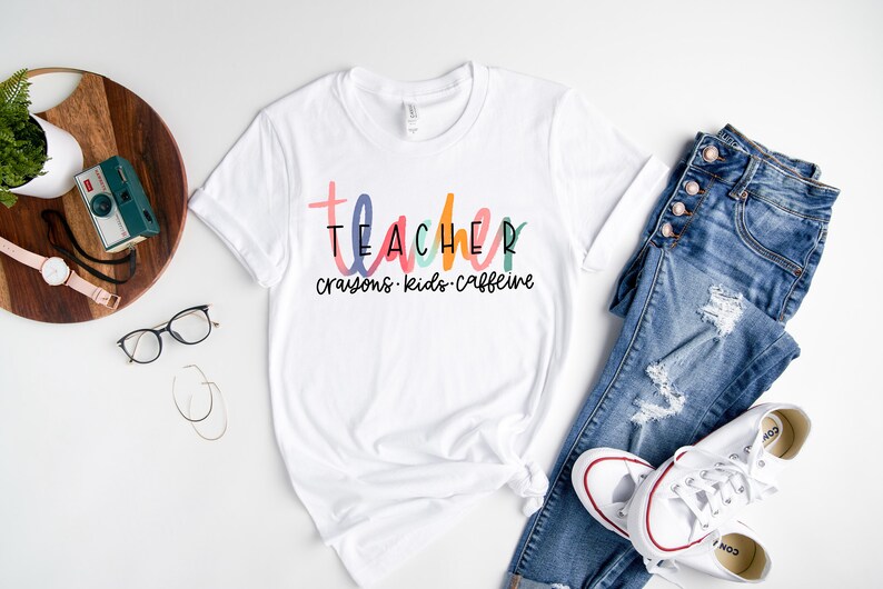 May include: White t-shirt with the word "TEACHER" in colorful letters, with "crayons, kids, caffeine" below. The shirt is paired with blue jeans and white sneakers.