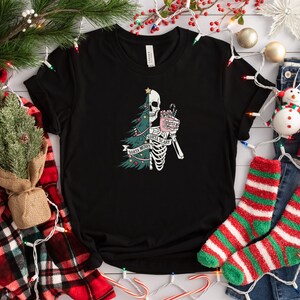 May include: A black short-sleeve t-shirt featuring a graphic print of a split figure that is half a green Christmas tree and half a white skeleton. The skeleton holds a pink mug with a candy cane. The text reads Santa Merry Santa Scary.