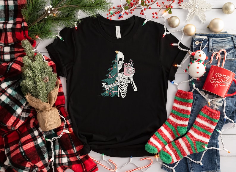 May include: A black short-sleeve t-shirt featuring a graphic print of a split figure that is half a green Christmas tree and half a white skeleton. The skeleton holds a pink mug with a candy cane. The text reads Santa Merry Santa Scary.