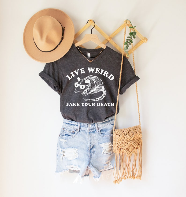 Puede incluir: Una camiseta gris oscuro con el texto "LIVE WEIRD FAKE YOUR DEATH" y un gr&aacute;fico de una zarig&uuml;eya con gafas. El conjunto incluye pantalones cortos de mezclilla, un sombrero beige y un bolso de macram&eacute;.