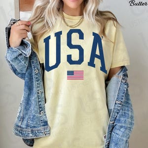 May include: A pale yellow t-shirt featuring the word "USA" in navy blue, with a small American flag beneath. The shirt is paired with a denim jacket and a gold chain necklace.