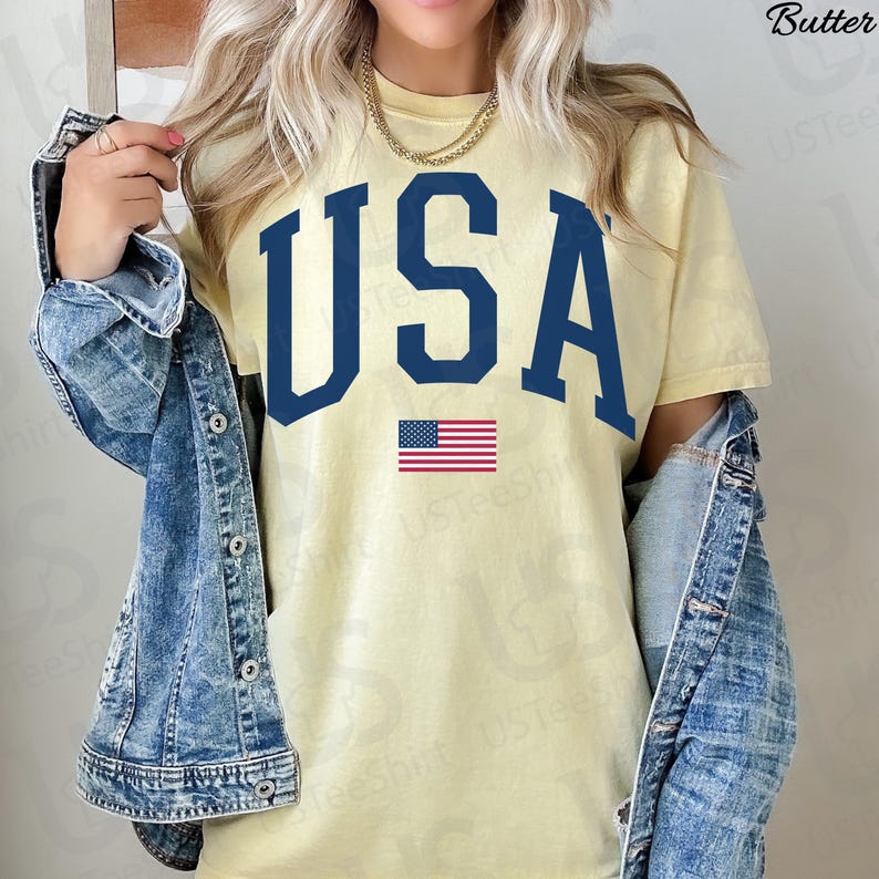 May include: A pale yellow t-shirt featuring the word "USA" in navy blue, with a small American flag beneath. The shirt is paired with a denim jacket and a gold chain necklace.
