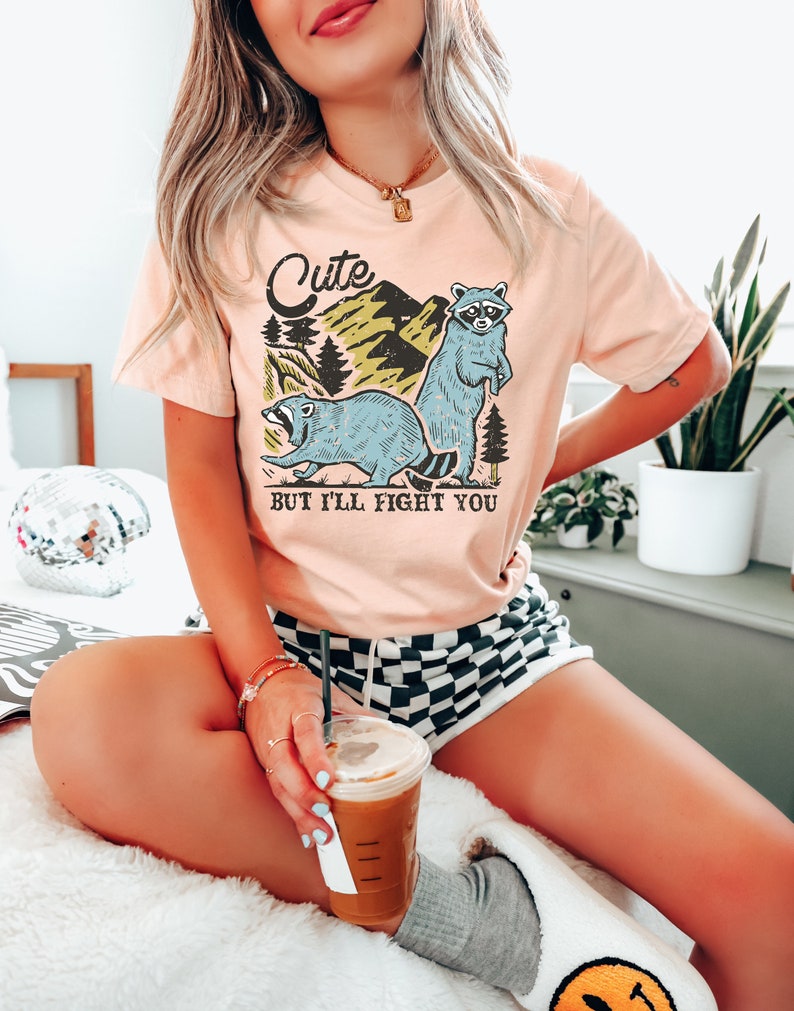 May include: Peach-colored t-shirt with a graphic of two blue raccoons and the text "Cute But I'll Fight You." The design includes mountains and trees. The shirt is a short-sleeved, casual top.