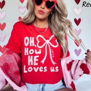 May include: A red long-sleeve shirt with the text "OH, HOW HE LOVES US" in white. The shirt features a large white bow design. The person is wearing heart-shaped sunglasses and a pink and red sweater.