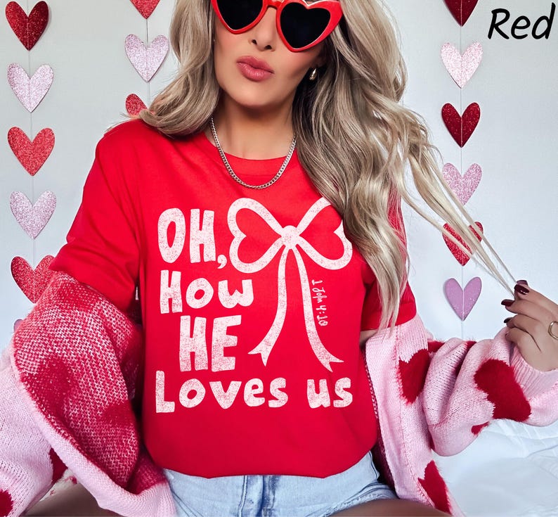 May include: A red long-sleeve shirt with the text "OH, HOW HE LOVES US" in white. The shirt features a large white bow design. The person is wearing heart-shaped sunglasses and a pink and red sweater.