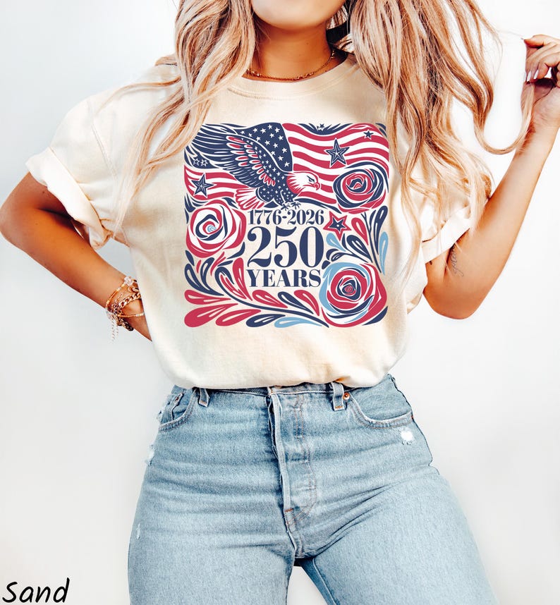 K&ouml;nnte beinhalten: Sandfarbenes T-Shirt mit patriotischem Design. Die Grafik zeigt eine amerikanische Flagge, einen Adler, Rosen und den Text "1776-2026 250 YEARS" in Rot, Wei&szlig; und Blau. Das Shirt ist ein l&auml;ssiger Stil.