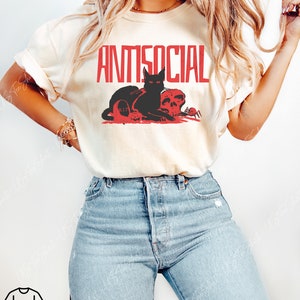 K&ouml;nnte beinhalten: Ein cremefarbenes Kurzarm-T-Shirt mit dem roten Schriftzug ANTISOCIAL &uuml;ber einer Grafik, die eine schwarze Katze, einen Totenkopf, einen Grabstein mit der Aufschrift R.I.P. und eine kleine Spinne zeigt. Dazu eine hellblaue Jeans.