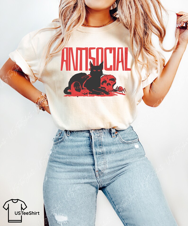 K&ouml;nnte beinhalten: Ein cremefarbenes Kurzarm-T-Shirt mit dem roten Schriftzug ANTISOCIAL &uuml;ber einer Grafik, die eine schwarze Katze, einen Totenkopf, einen Grabstein mit der Aufschrift R.I.P. und eine kleine Spinne zeigt. Dazu eine hellblaue Jeans.