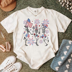 May include: Ivory-colored t-shirt featuring a festive design of dancing skeletons celebrating Independence Day. The skeletons are adorned with patriotic elements like the American flag and fireworks. The shirt is paired with white sneakers and accessories.